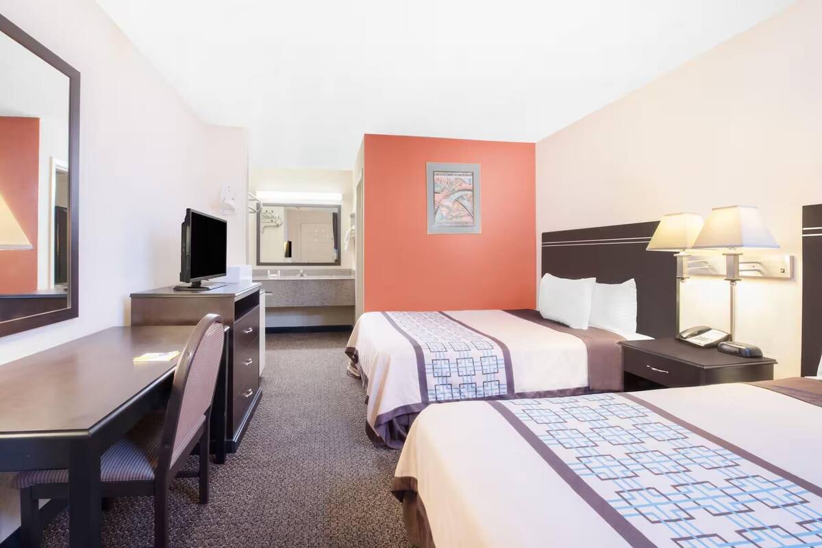 A hotel room with a desk, two beds, a television, a mirror, and a painting on the wall.