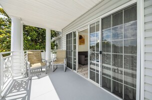 A covered porch with two wicker chairs and a table, a sliding glass door leading to an outdoor seating area, and a white exterior wall.