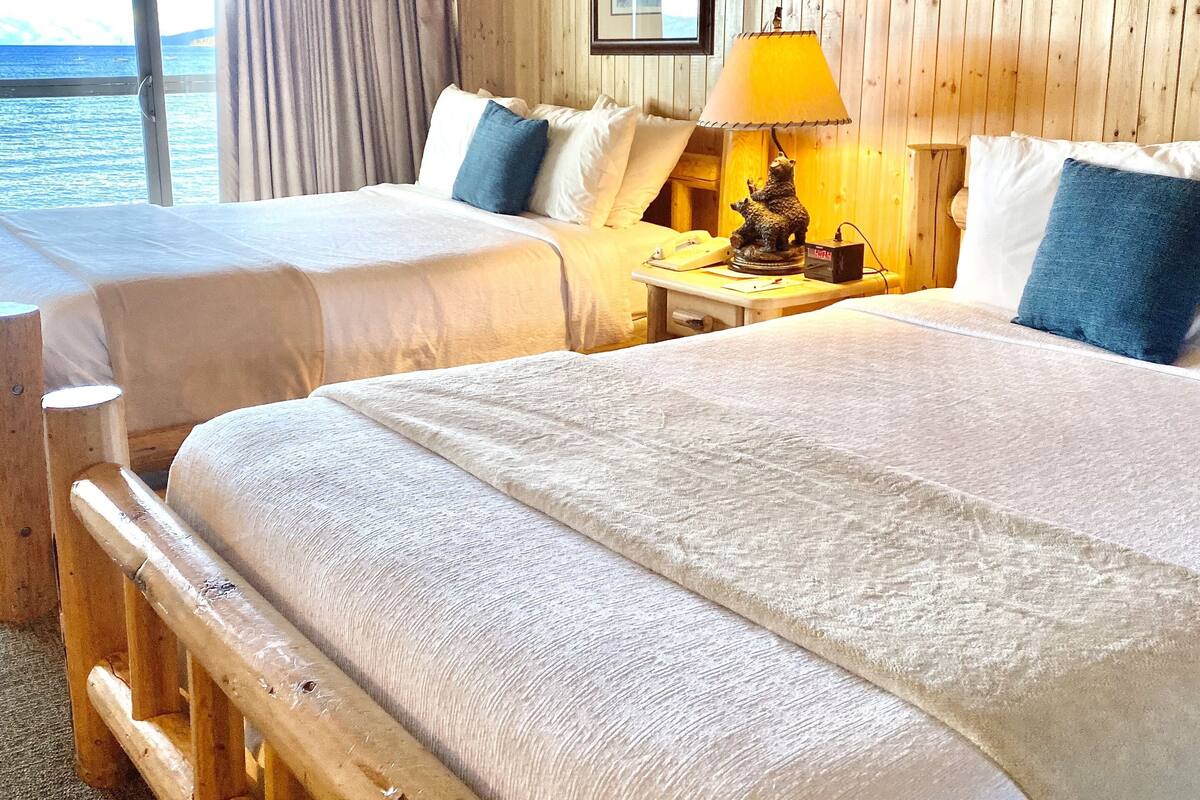 A hotel room with two beds, wooden walls, a view of the sea, and a lamp on the nightstand.