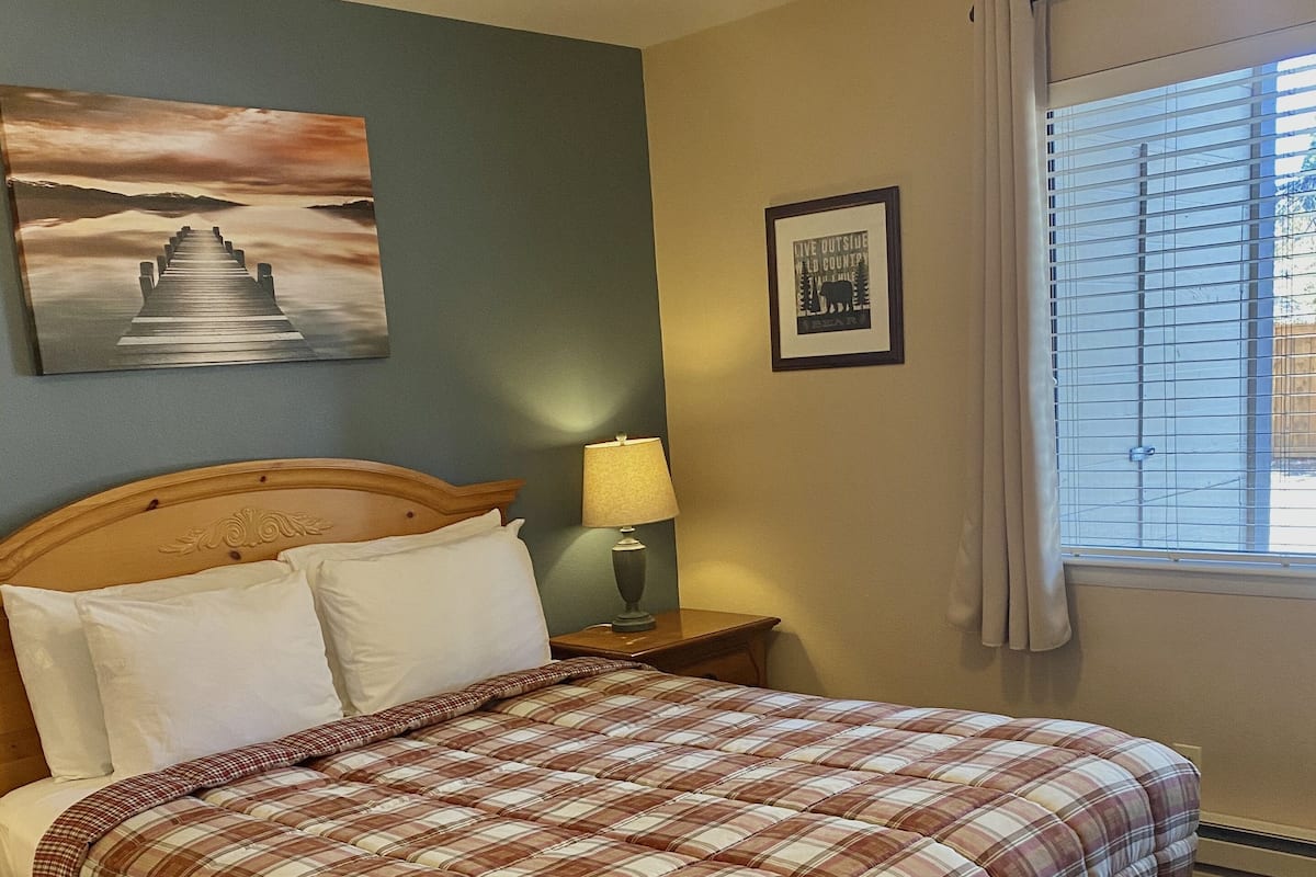 A neatly made bed with a plaid comforter, a wooden headboard, a nightstand with a lamp, a framed picture on the wall, and a window with blinds.