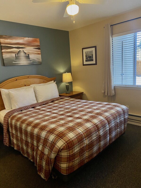 A neatly made bed with a plaid comforter, a wooden headboard, a nightstand with a lamp, a framed picture on the wall, and a window with blinds. - Tahoe Lakeshore Lodge & Spa (South Lake Tahoe)