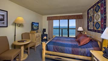A cozy room with a bed, a desk, a chair, a small table, a lamp, a TV, and a fireplace.