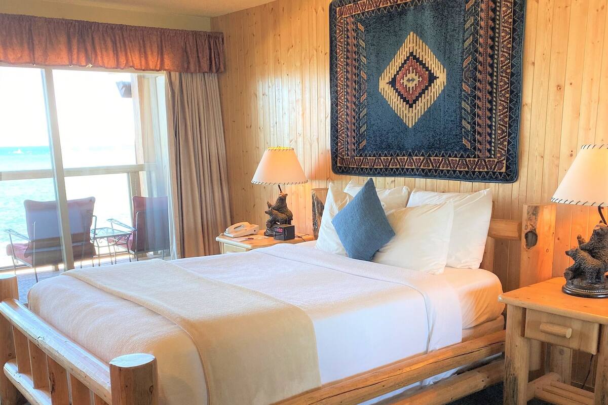 A wooden-framed bed with white linens, a patterned rug above it, and a view of the sea through a large window.