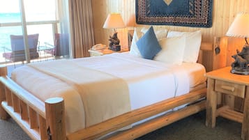 A wooden-framed bed with white linens, a patterned rug above it, and a view of the sea through a large window.