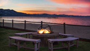A wooden deck with a stone fire pit and benches overlooking a lake with mountains in the background.