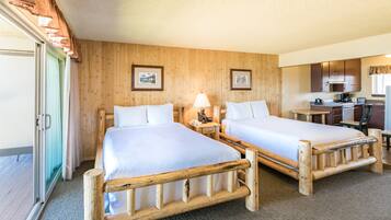 A room with two wooden beds, a small desk, a chair, a lamp, and a kitchenette.