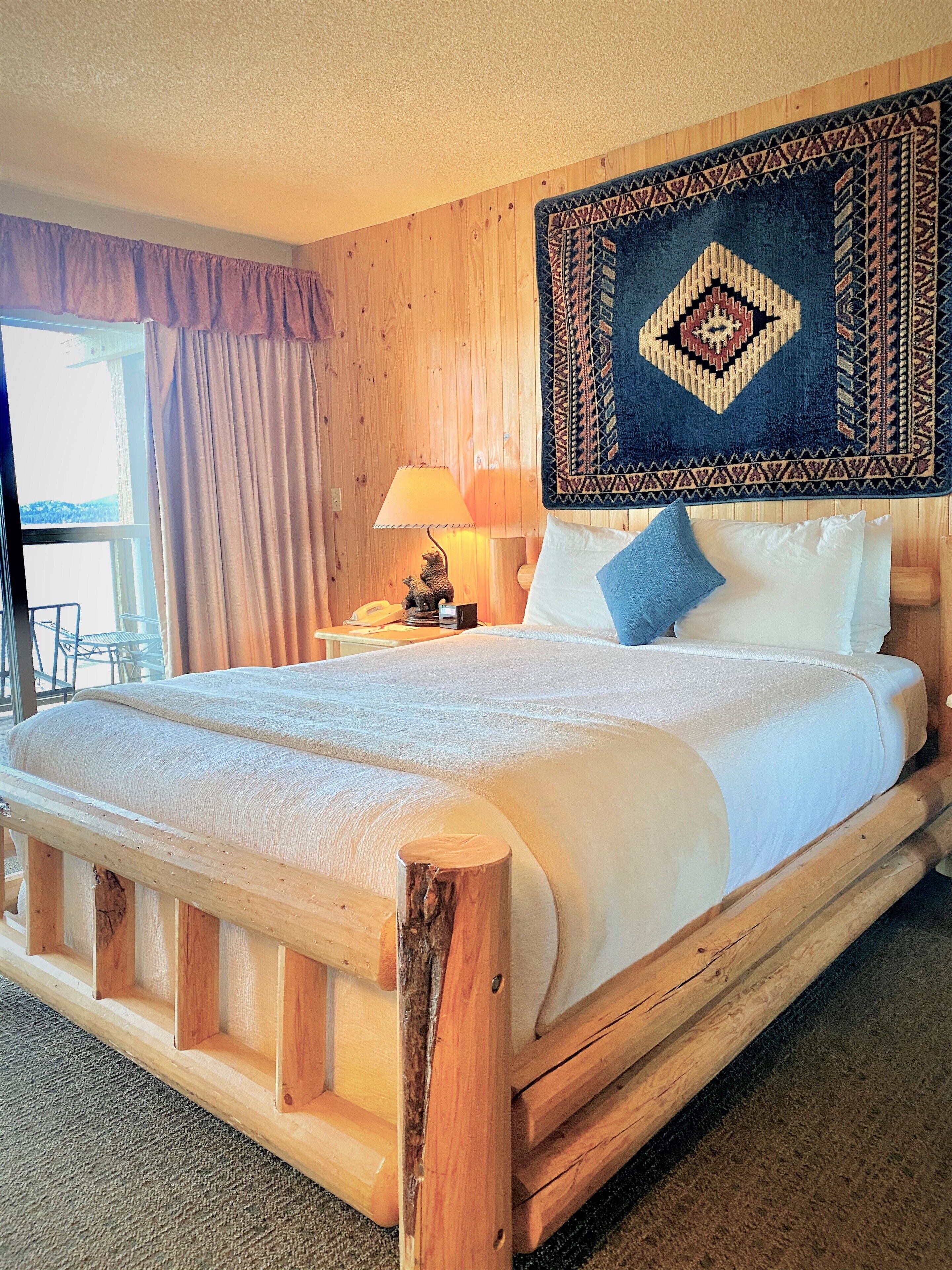 Una cama de madera con cabecera, una mesita de noche con lámpara y una alfombra estampada.