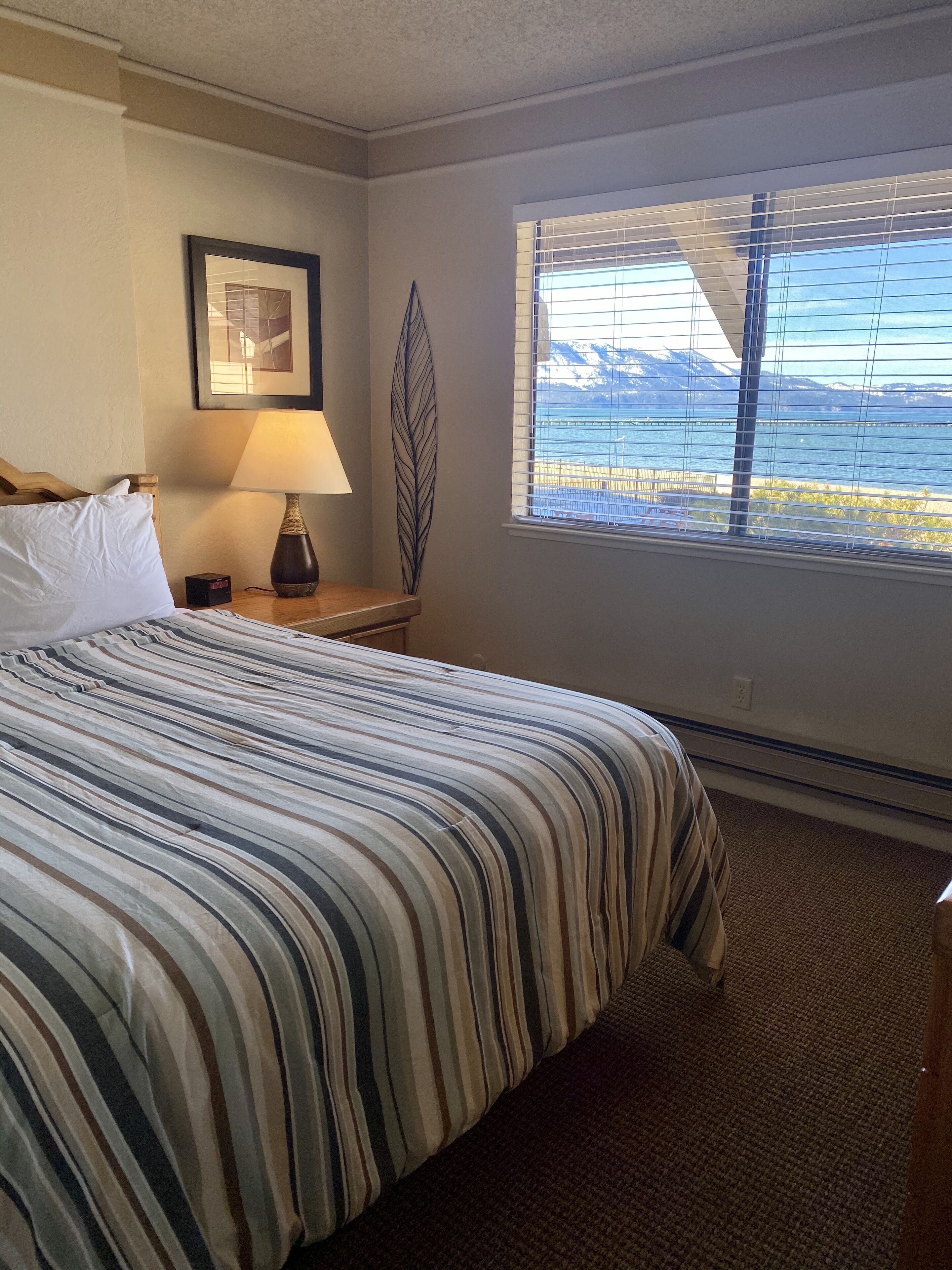 Habitación de hotel con una cama grande, una mesita de noche con lámpara, un cuadro enmarcado en la pared y una ventana con vistas a montañas y agua.