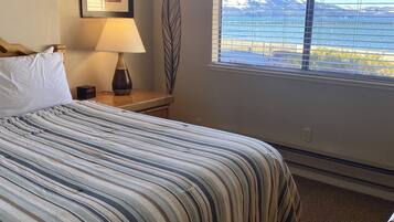 A hotel room with a large bed, a nightstand with a lamp, a framed picture on the wall, and a window with a view of mountains and water.