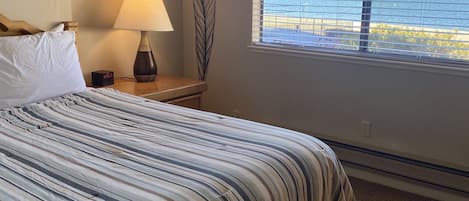 Habitación de hotel con una cama grande, una mesita de noche con lámpara, un cuadro enmarcado en la pared y una ventana con vistas a montañas y agua.