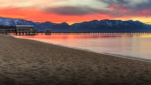 A beach with a pier extending into calm waters, mountains in the background, and a building with a flat roof on the left.