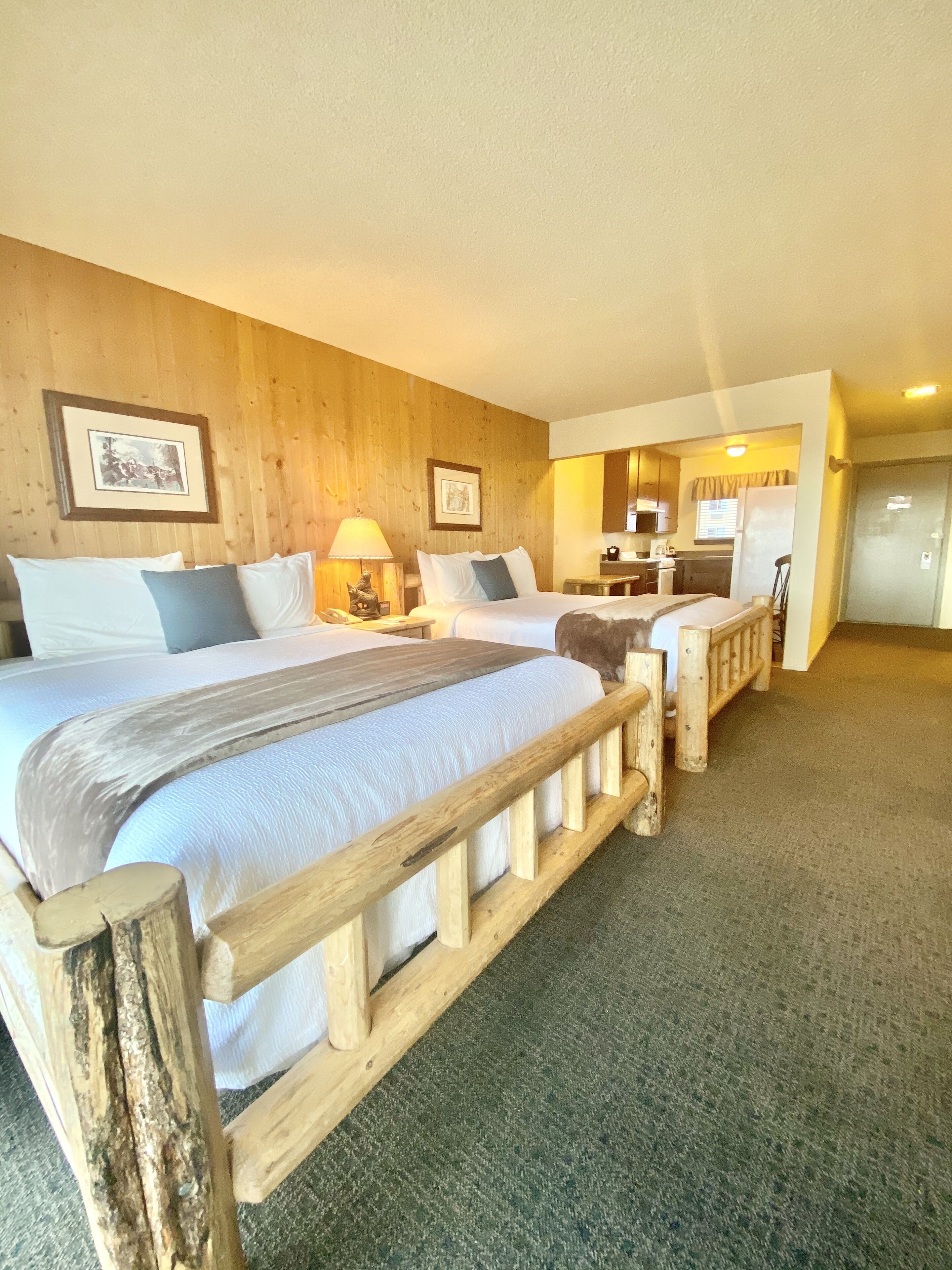 Habitación de hotel con dos camas, cabeceros de madera, una kitchenette pequeña y un baño con ducha.