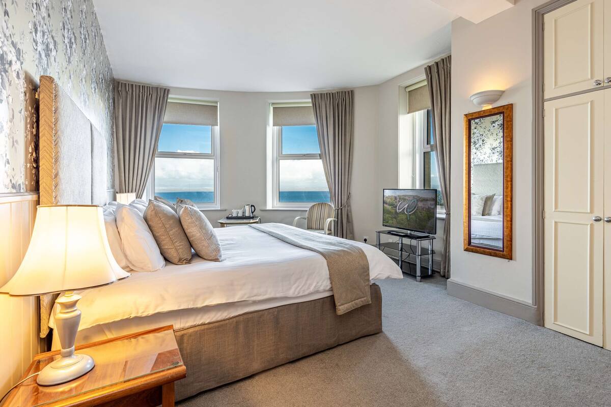 A hotel room with a large bed, a desk with a chair, a TV, and a view of the sea.