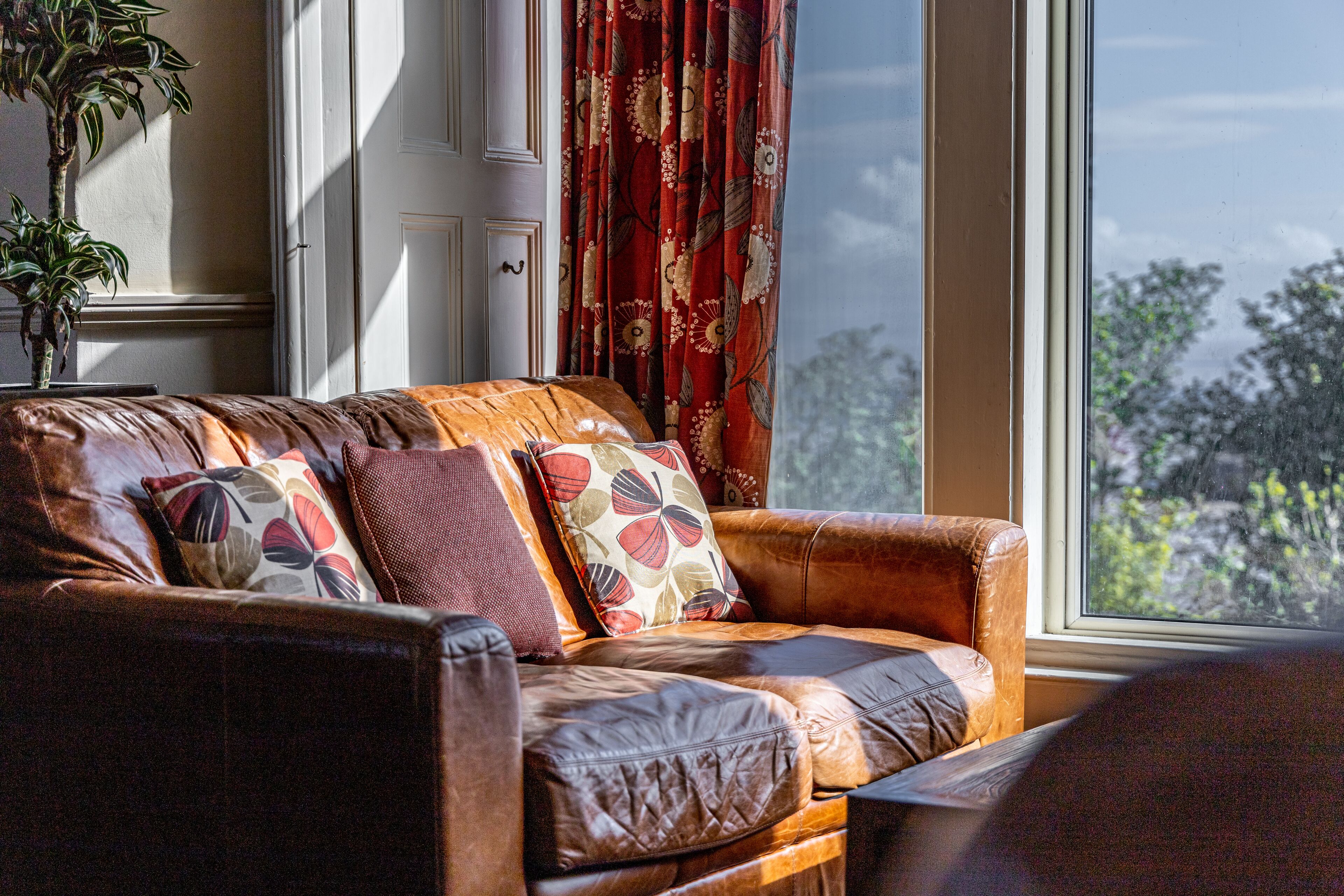 A leather sofa with floral cushions, positioned near a window with a view of trees.