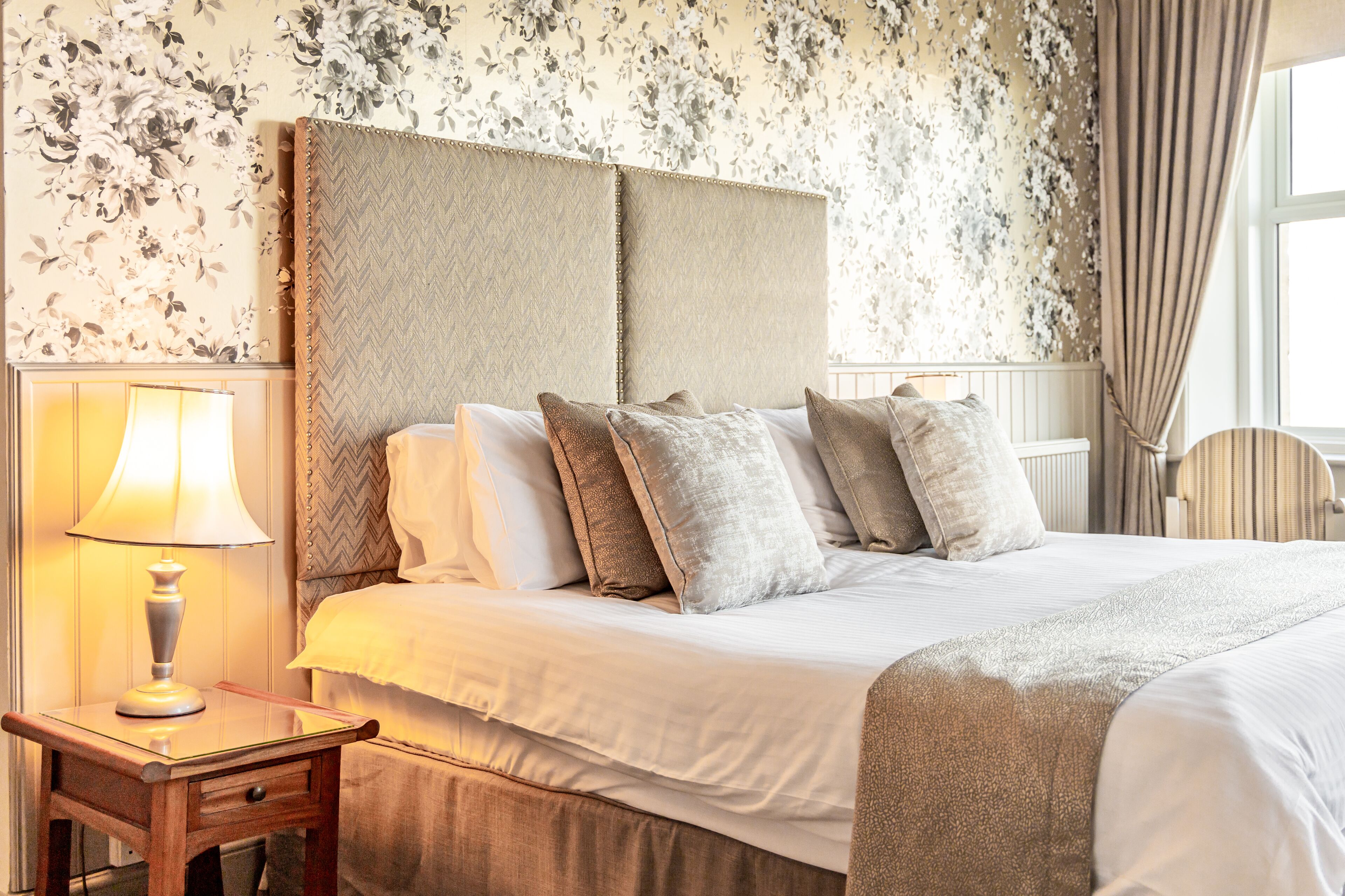 A neatly made bed with a headboard, a bedside table with a lamp, and floral wallpaper.