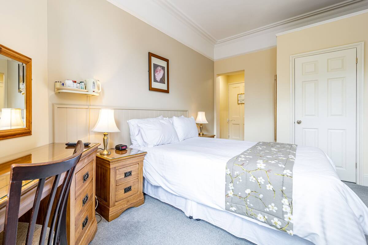 A hotel room with a bed, a desk, a chair, a bedside table with lamps, a wall-mounted shelf with toiletries, and a door.
