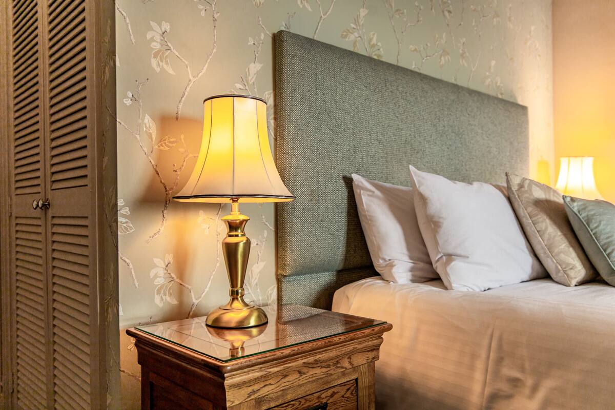 A neatly made bed with pillows, a bedside lamp on a wooden nightstand, and floral wallpaper.