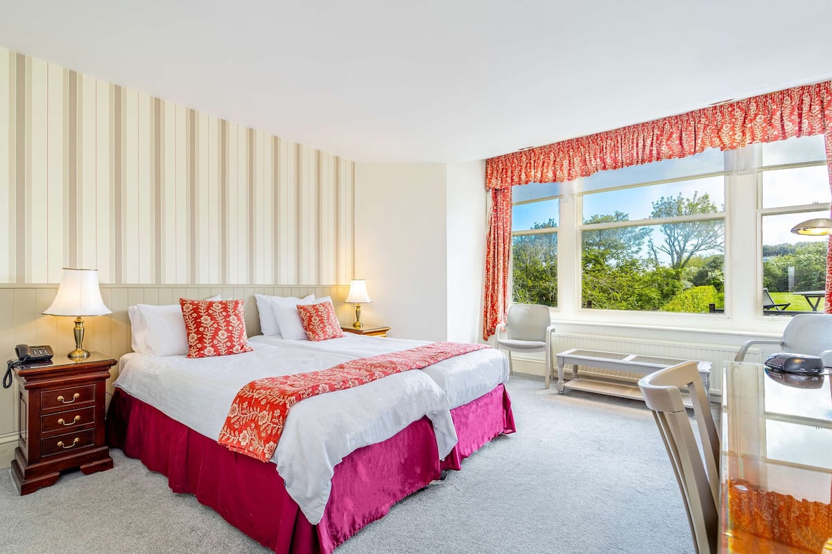 A hotel room with a large bed, two bedside tables with lamps, a desk with a chair, and a window with a view of greenery.