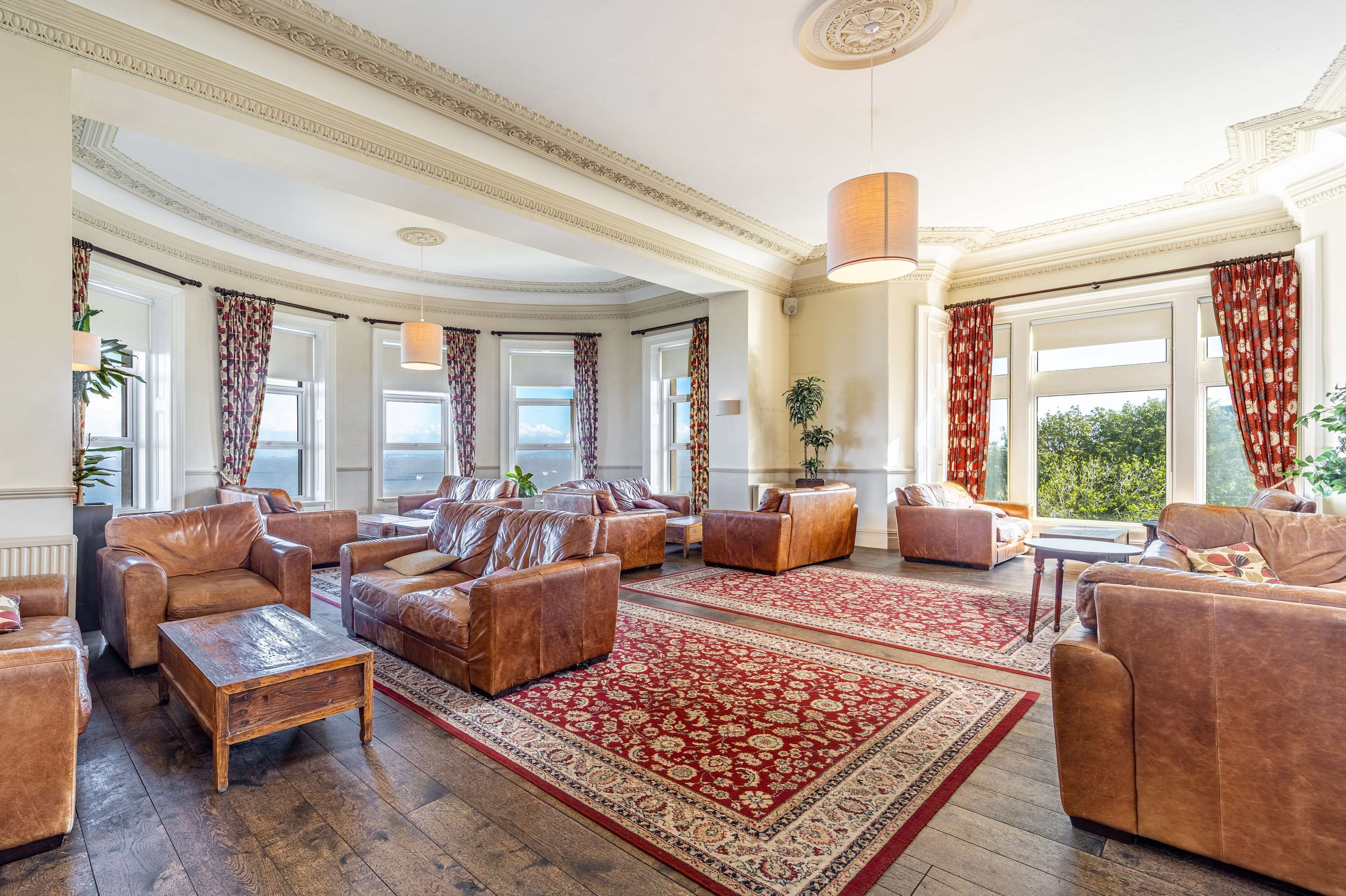 A spacious living room with leather sofas, a large red patterned rug, and large windows with patterned curtains.