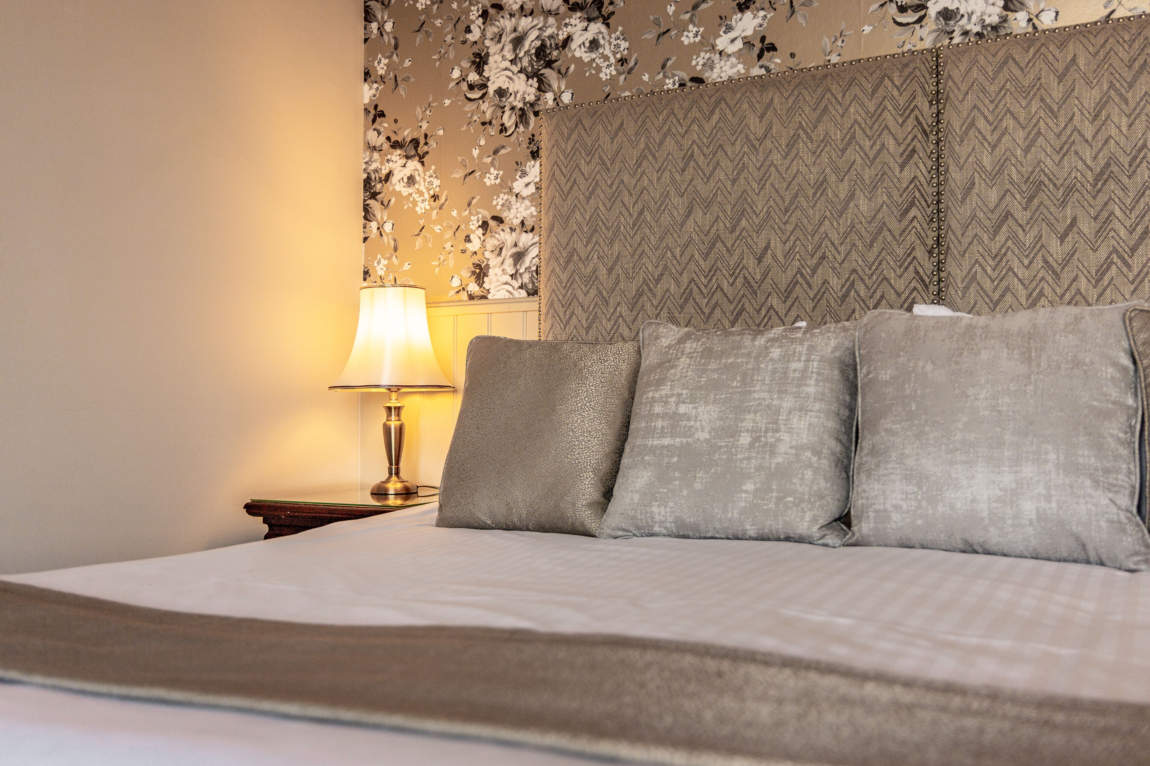 A neatly made bed with a patterned headboard, a bedside lamp, and a floral wallpaper design.