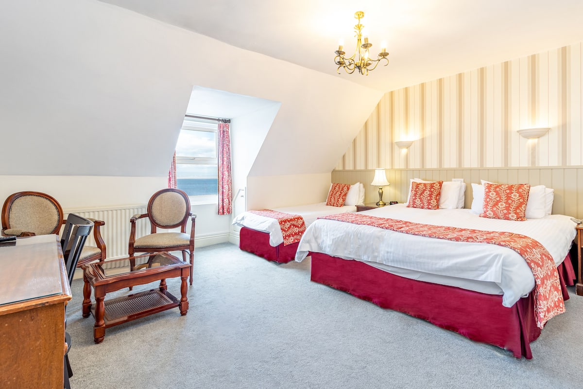 A hotel room with two beds, a desk, a chair, a chandelier, and a window with curtains.