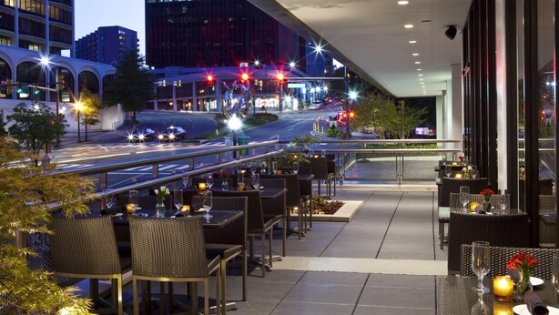 Un espacio exterior para comer con mesas y sillas, vista a una calle iluminada de noche y edificios modernos al fondo.