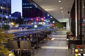 An outdoor dining area with tables and chairs, a view of a city street at night, and modern buildings in the background. - Hyatt Centric Arlington (Arlington)