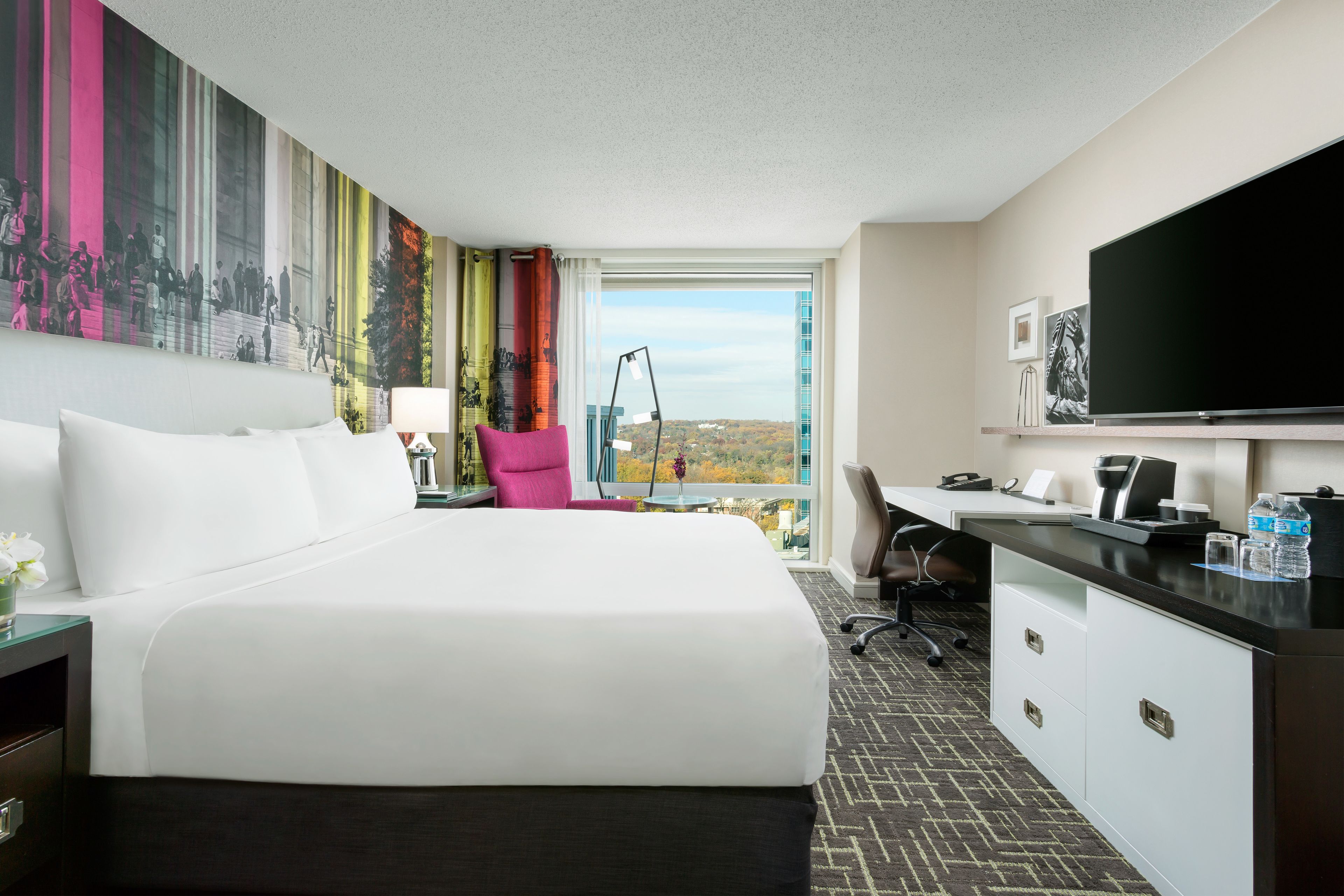 A hotel room with a large bed, a desk, a chair, a TV, and a view of the cityscape.