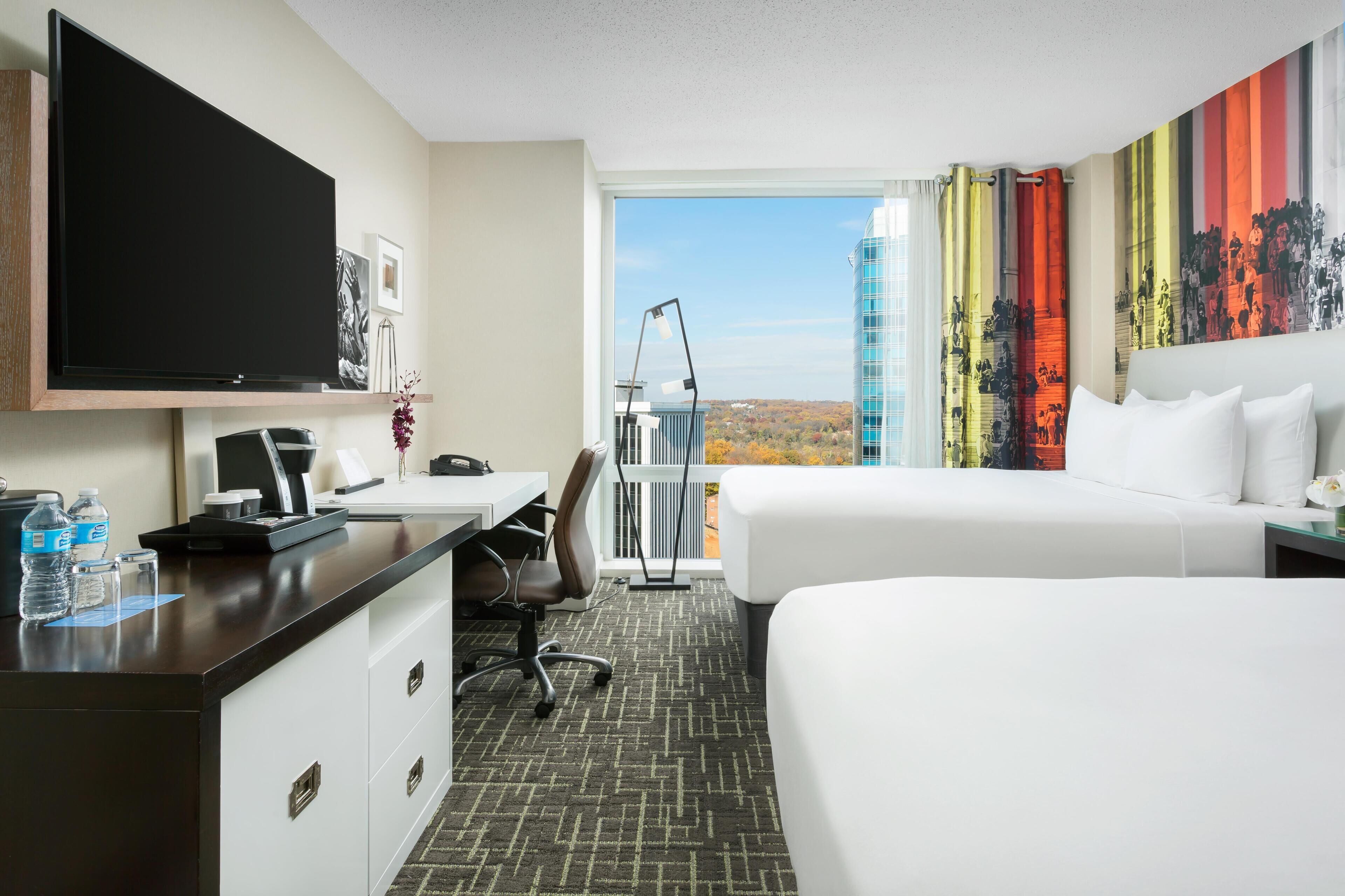 A hotel room with a bed, a desk with a flat-screen TV, a chair, a desk lamp, a water bottle, and a window with a cityscape view.