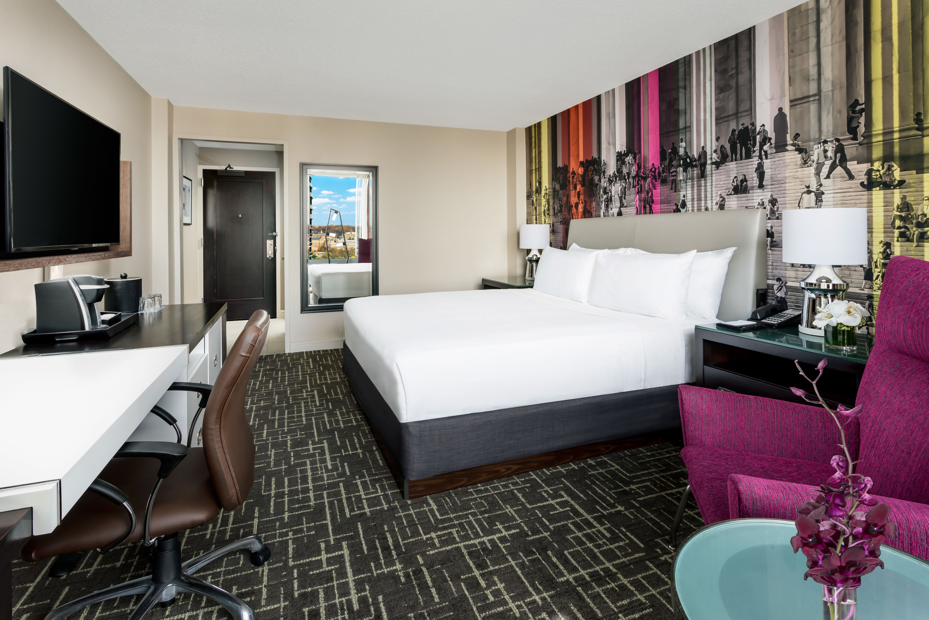 A hotel room with a bed, desk, chair, TV, and a painting on the wall.