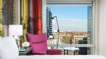 Habitación de hotel con una cama, un sillón rosa, una mesita con un florero, una lámpara y vistas a un paisaje urbano a través de amplias ventanas.