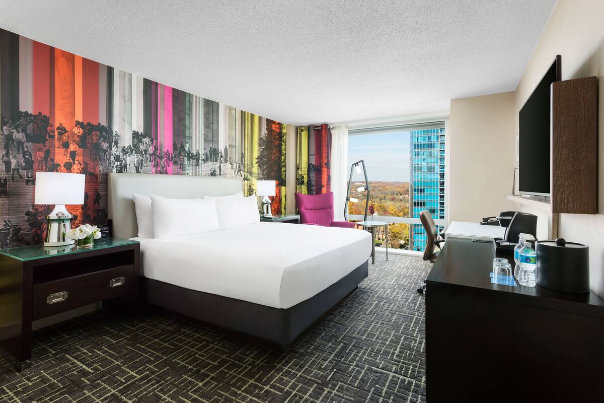 A hotel room with a large bed, a desk, a chair, a TV, and a window with a view of a building.