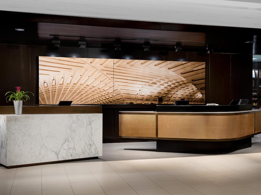 A reception desk with a marble base and a wooden panel design, a potted plant, and a modern ceiling with wooden slats.