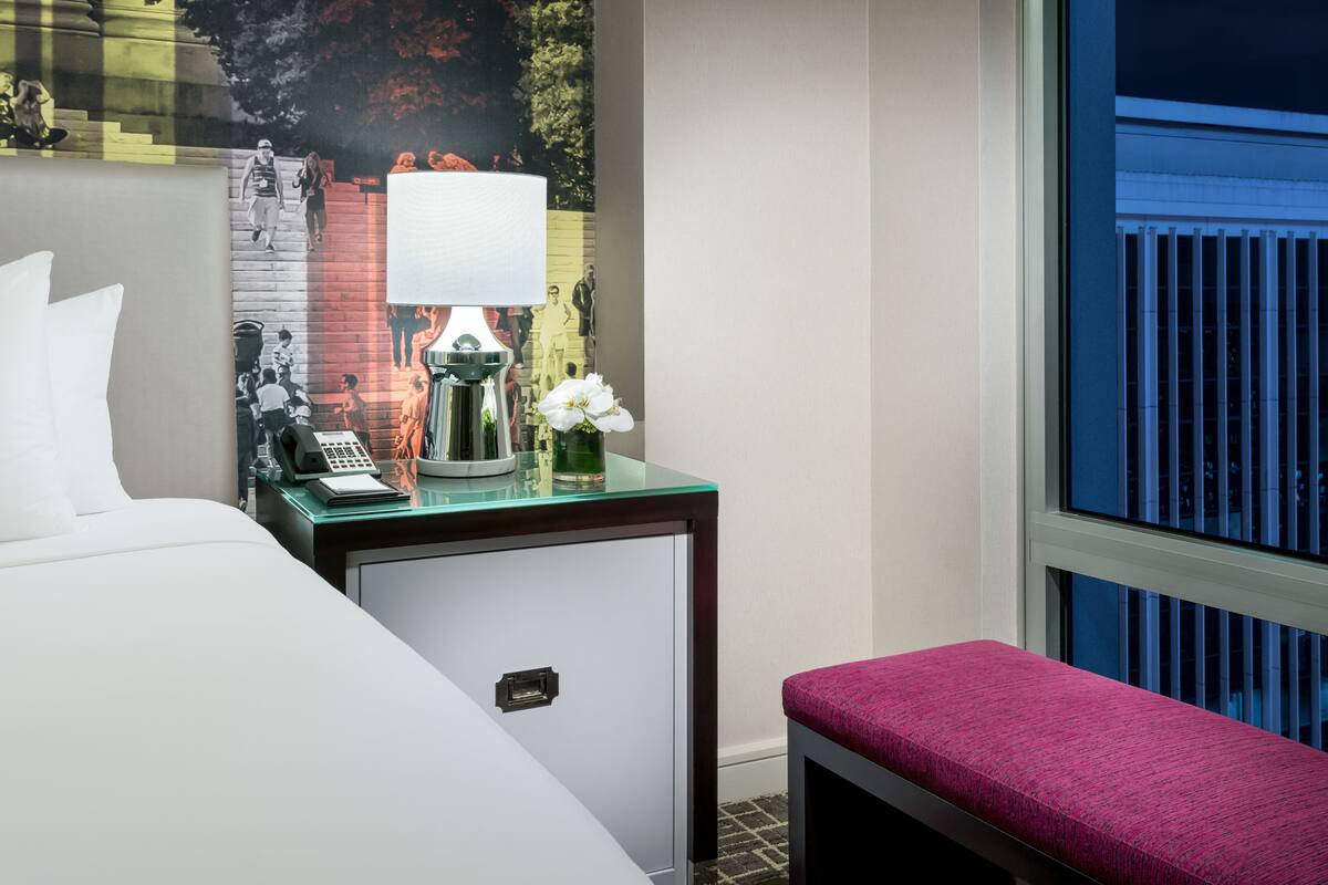 A hotel room with a bed, a nightstand with a lamp, a small cabinet, and a bench.