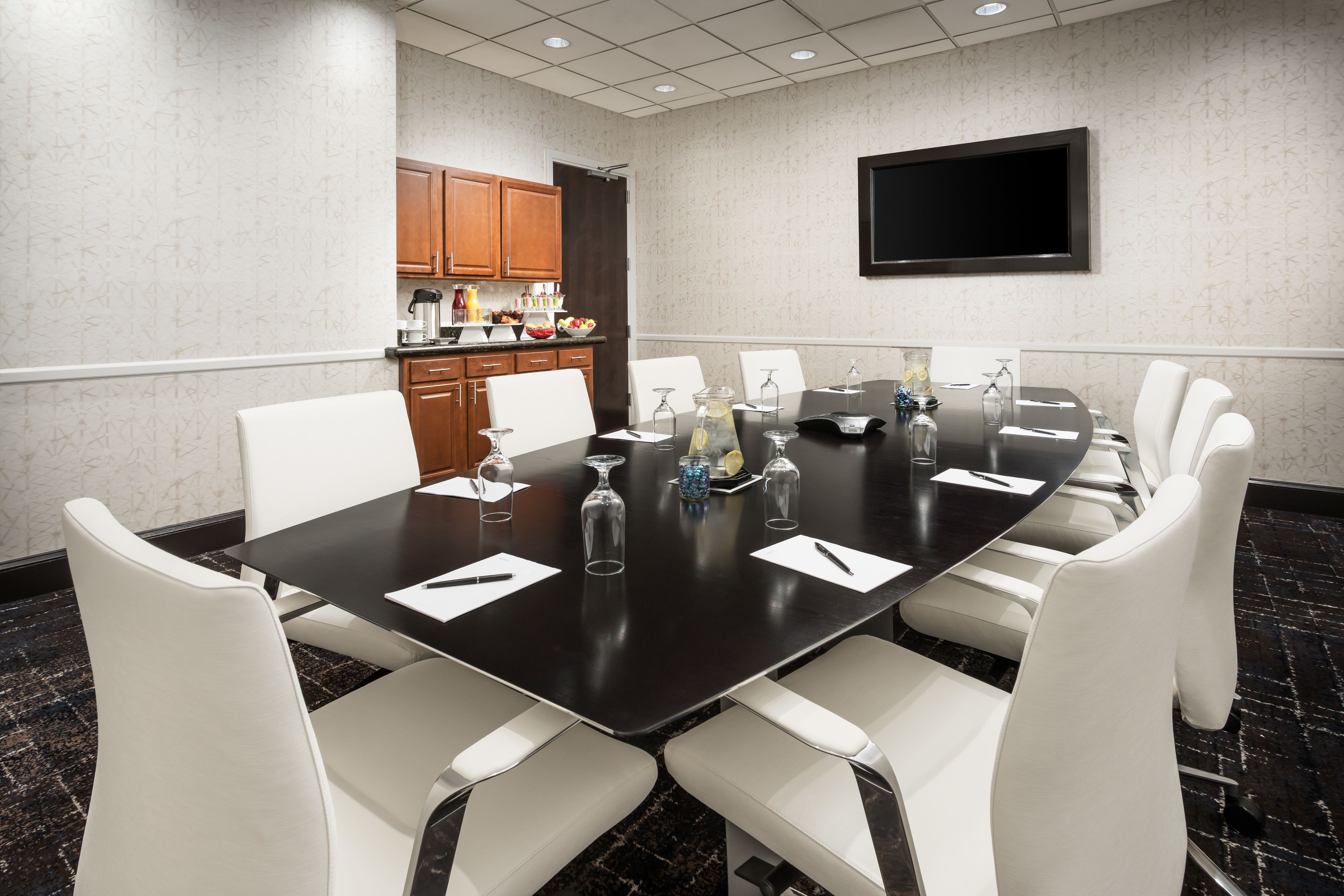A conference room with a long table, white chairs, a TV on the wall, and a kitchen area in the background.