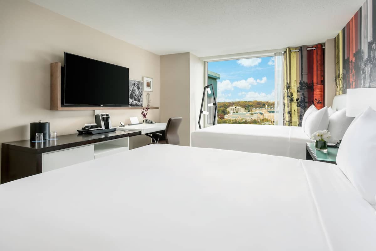 A hotel room with a bed, a desk with a flat-screen TV, a chair, a coffee maker, and a window with a view of buildings.