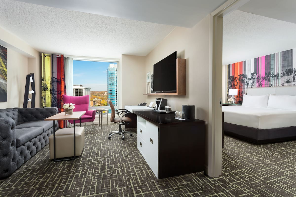 A hotel room with a gray sofa, a desk with a computer, a pink chair, a white bed, and a large window with a city view.