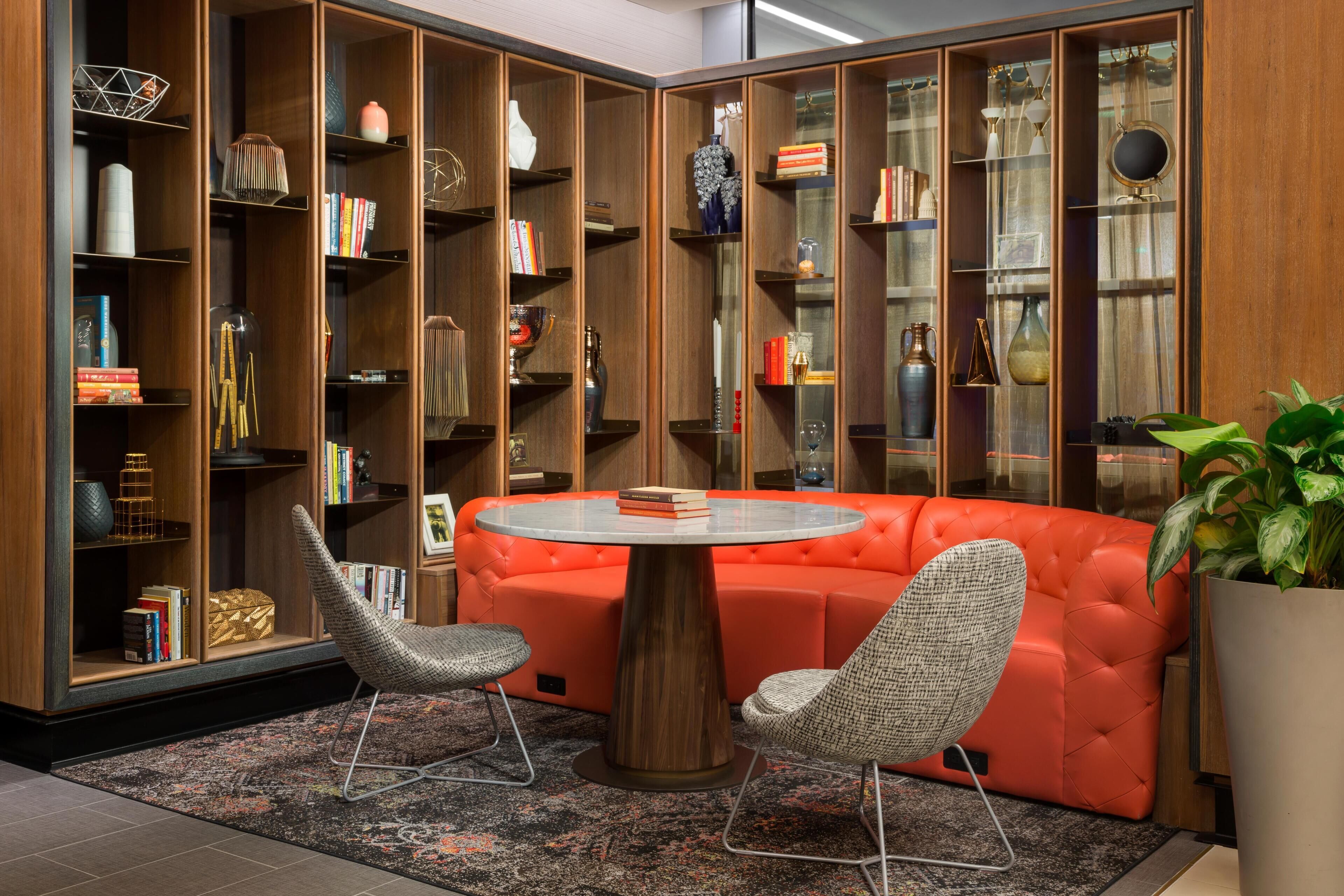Une pièce comprenant une table ronde, deux chaises, un canapé rouge et une grande bibliothèque remplie de livres et d’objets décoratifs.