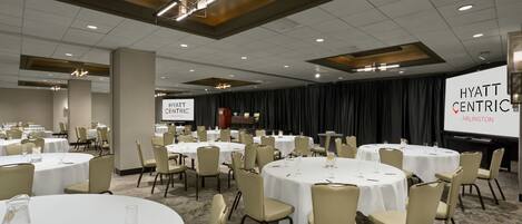 Un salón de conferencias con mesas y sillas redondas, una pantalla grande mostrando 'HYATT CENTRIC ARLINGTON' y un escenario con cortinas.
