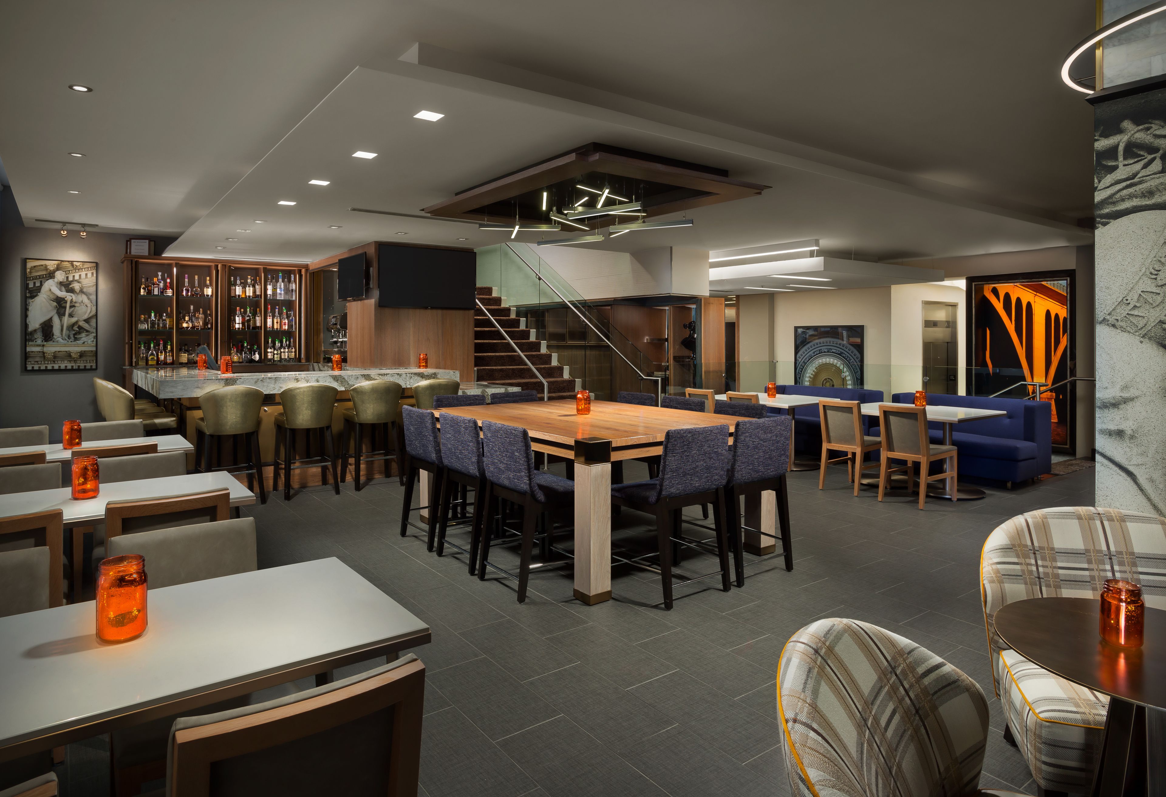 Un restaurante moderno con una mesa de madera central rodeada de sillas, una zona de bar con botellas y una escalera que lleva a un nivel superior.