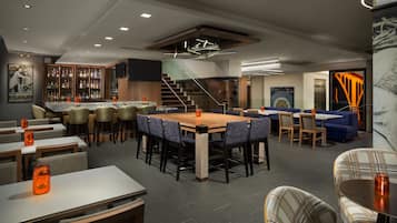 Un restaurante moderno con una mesa de madera central rodeada de sillas, una zona de bar con botellas y una escalera que lleva a un nivel superior.