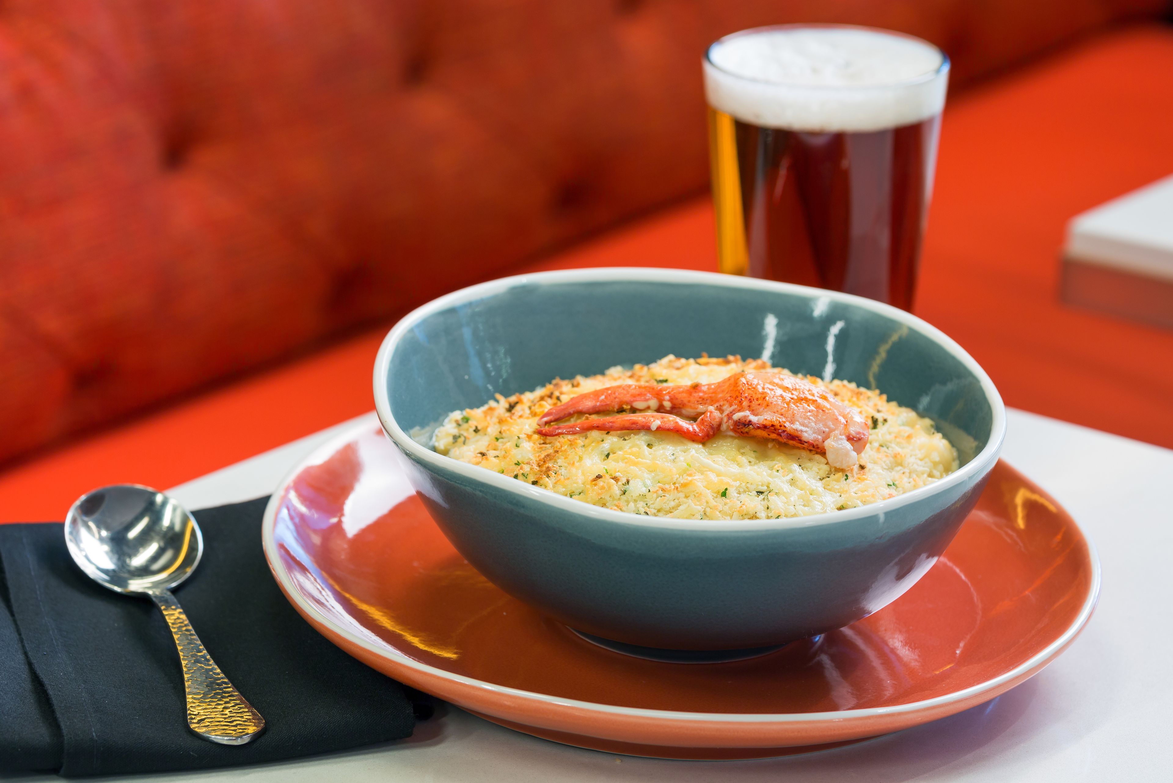 A bowl of food with a lobster on top, a spoon, and a glass of beer on a table.