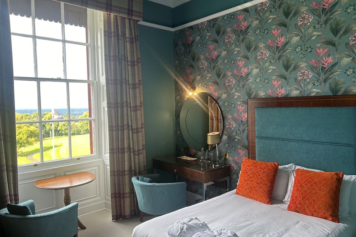 A hotel room with a bed, a desk, a chair, a round mirror, and a view of the outdoors.