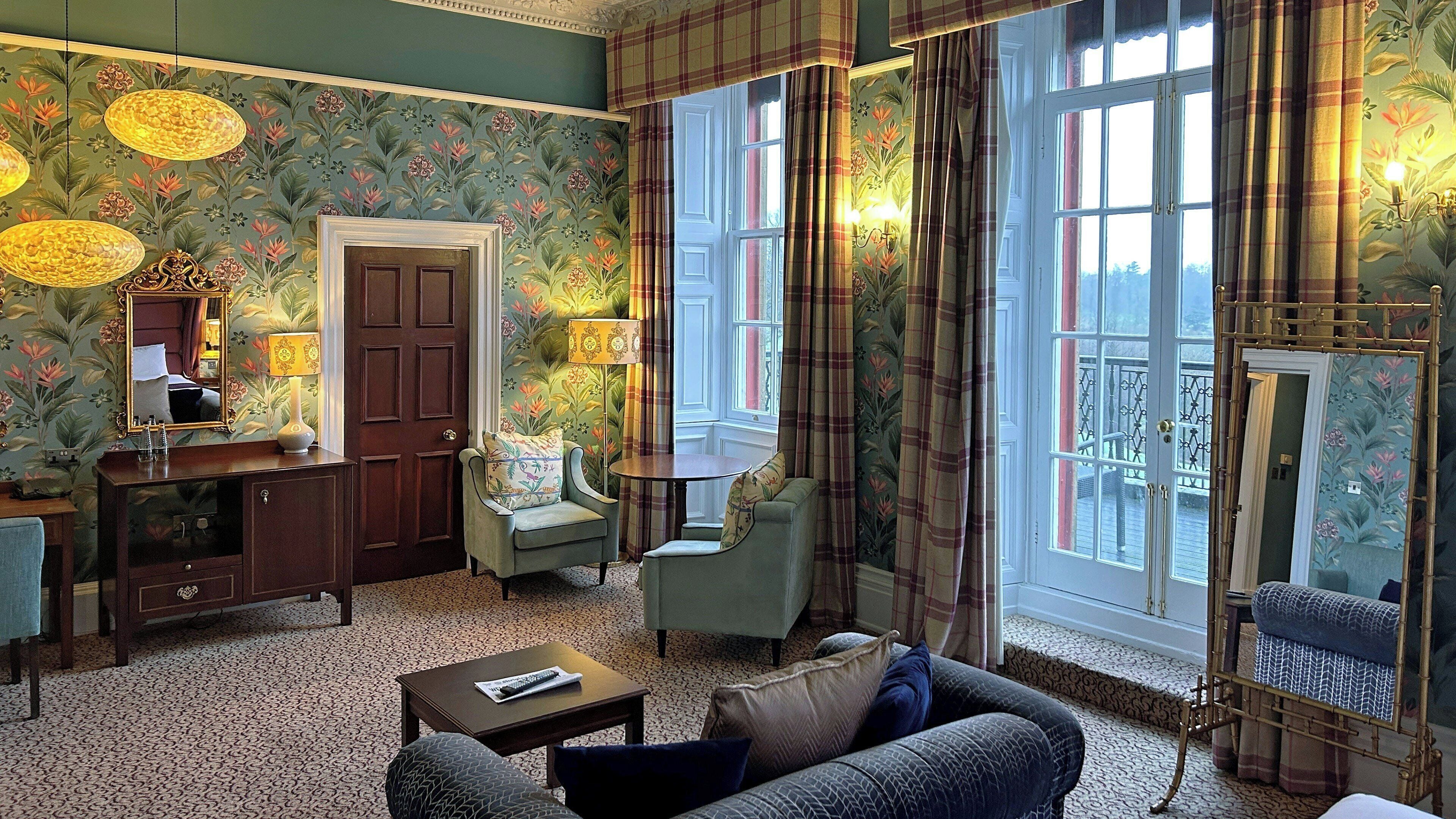 A room with floral wallpaper, a wooden dresser, a sofa, a table, and a window with curtains.