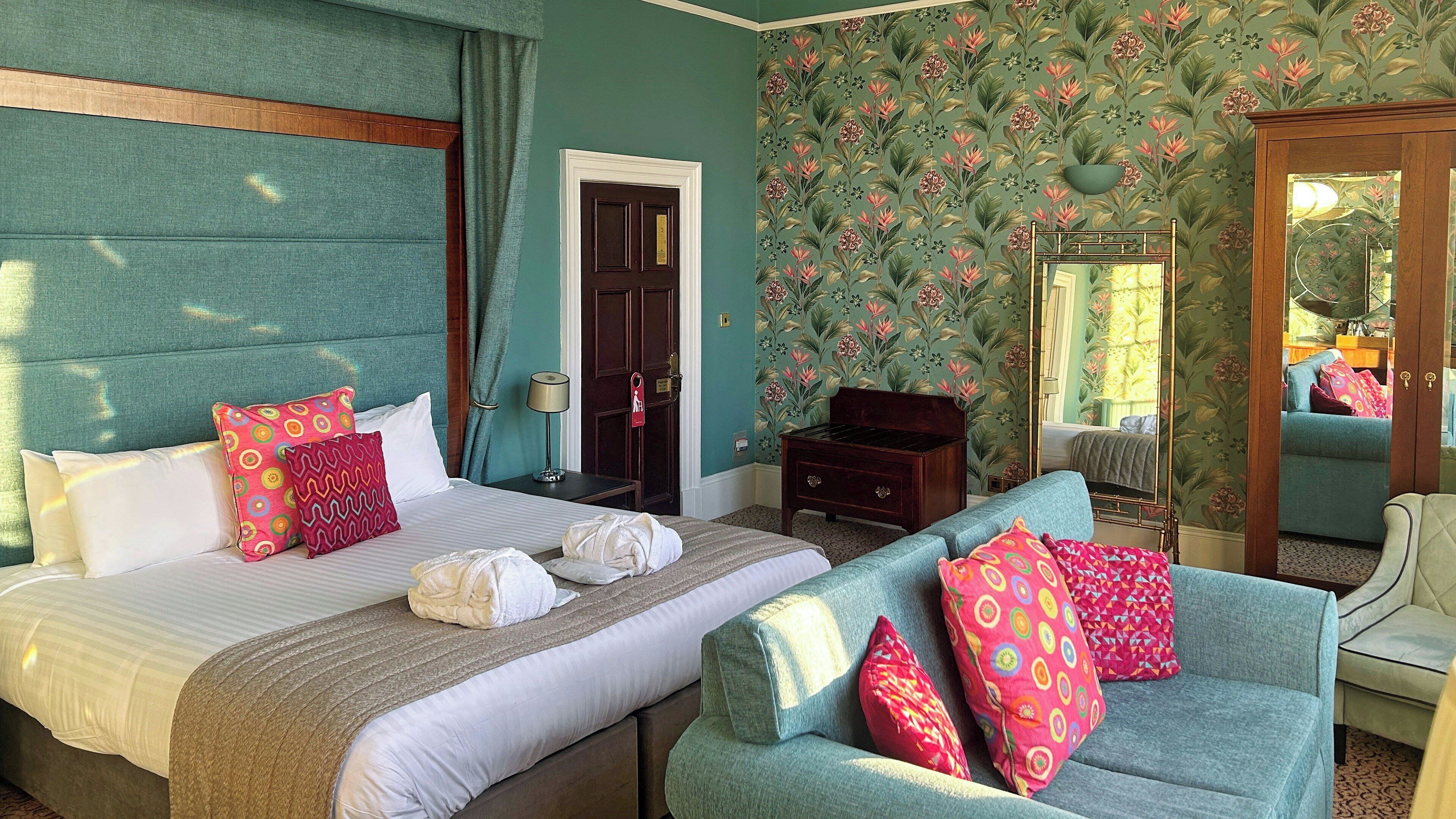 A hotel room with a bed, a sofa, a dresser, a mirror, and floral wallpaper.