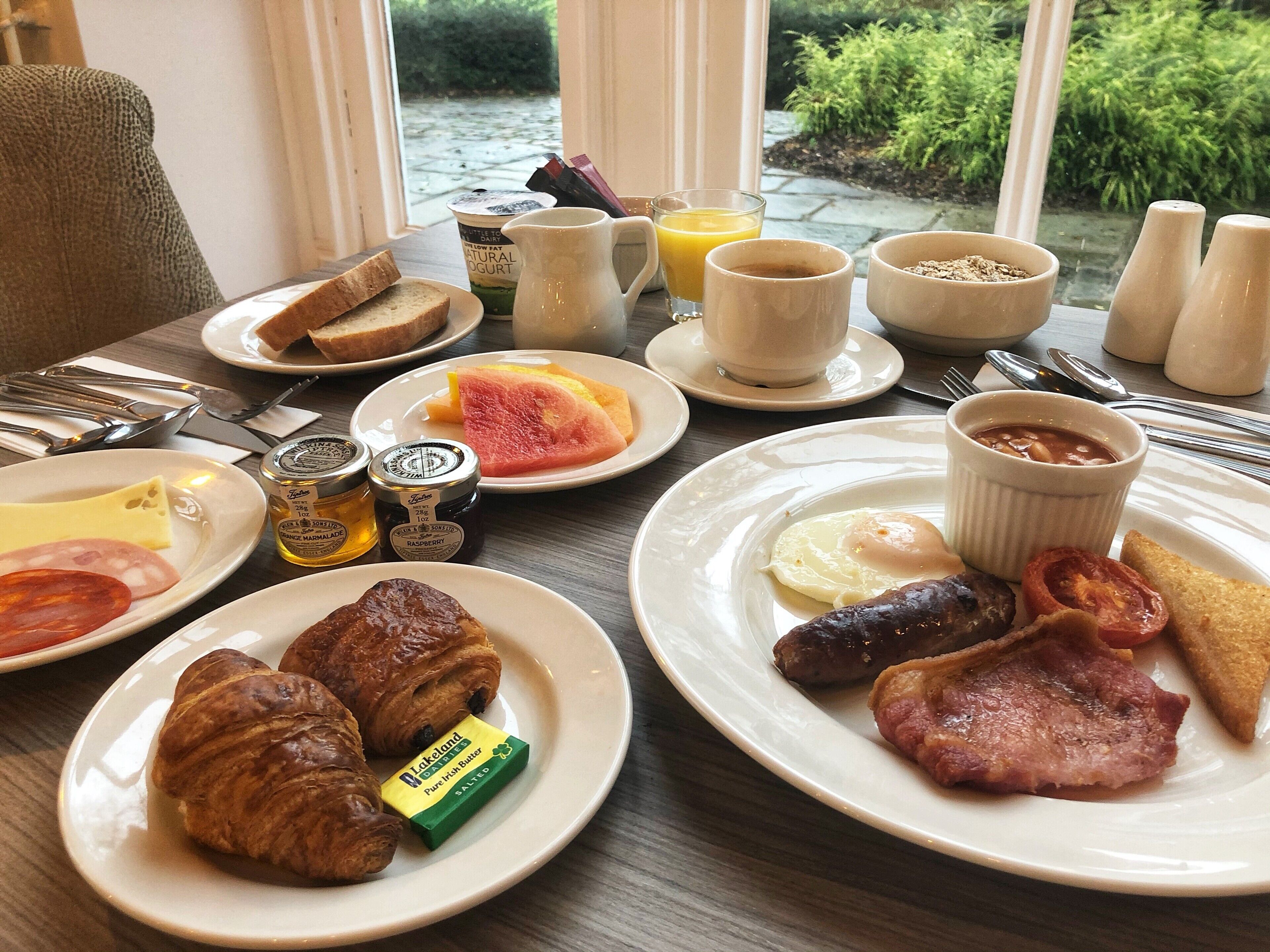 A breakfast table with croissants, ham, eggs, toast, jam, and a view of greenery outside.