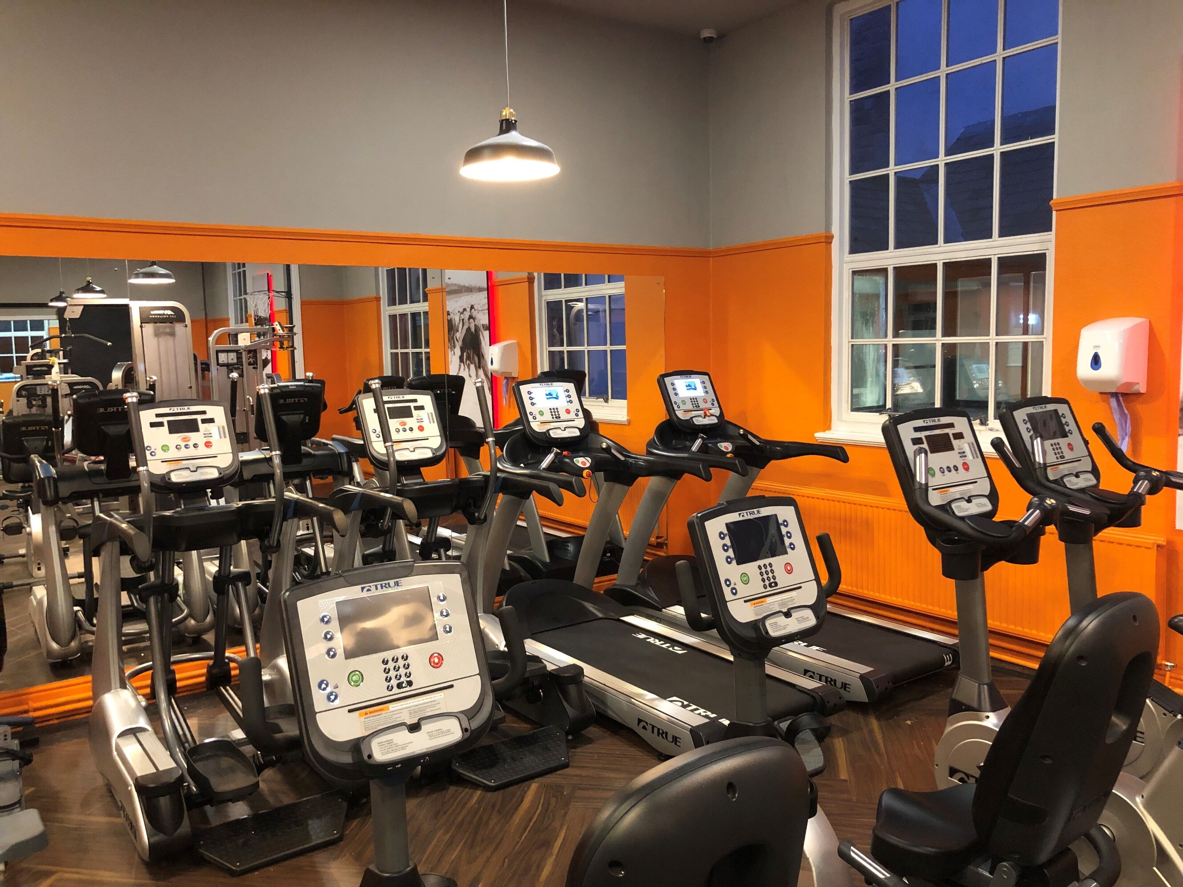 A gym with multiple treadmills and exercise machines, orange walls, and large windows.