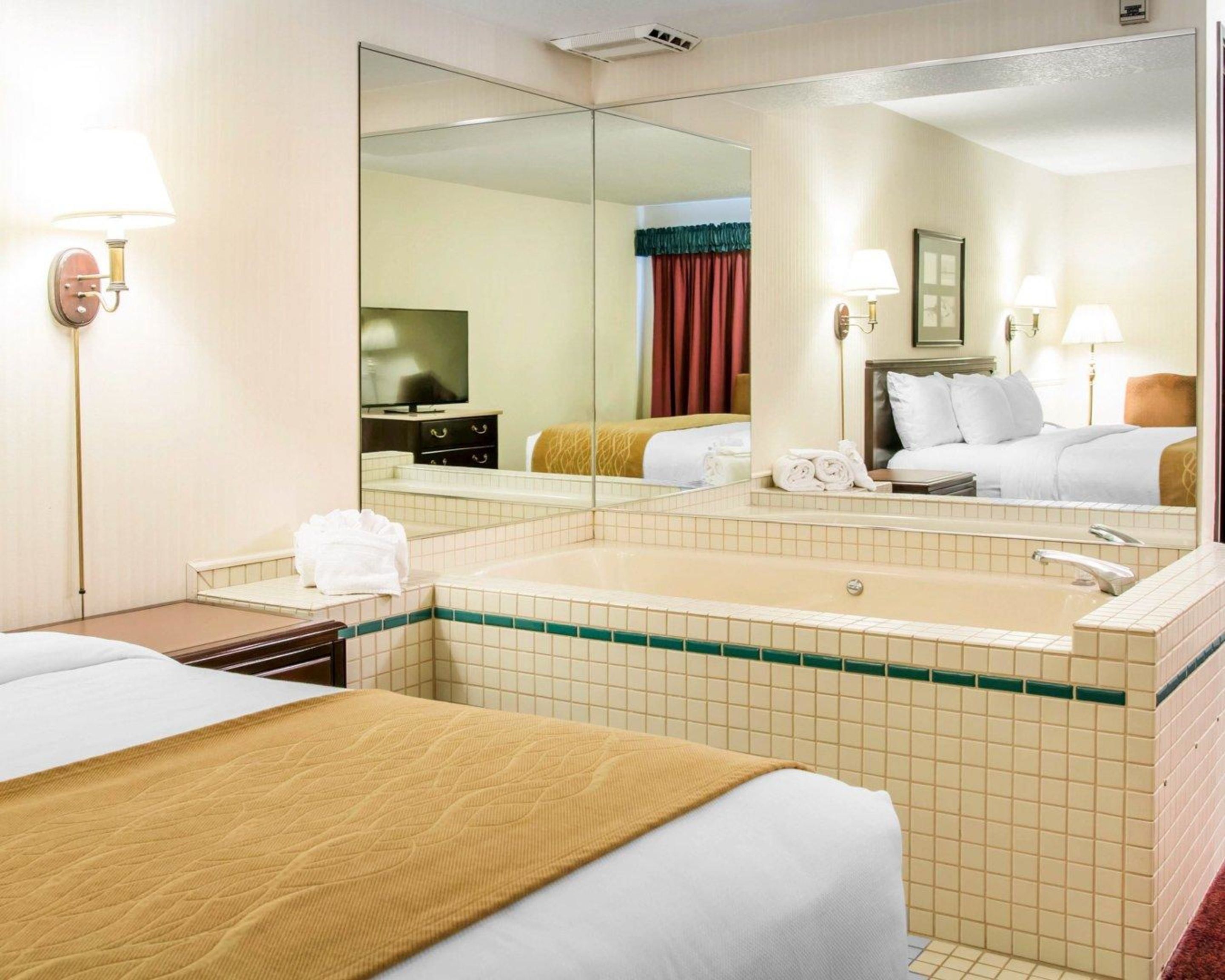 A hotel room with a large mirror, a bathtub, a bed with a yellow blanket, and a television on a dresser.