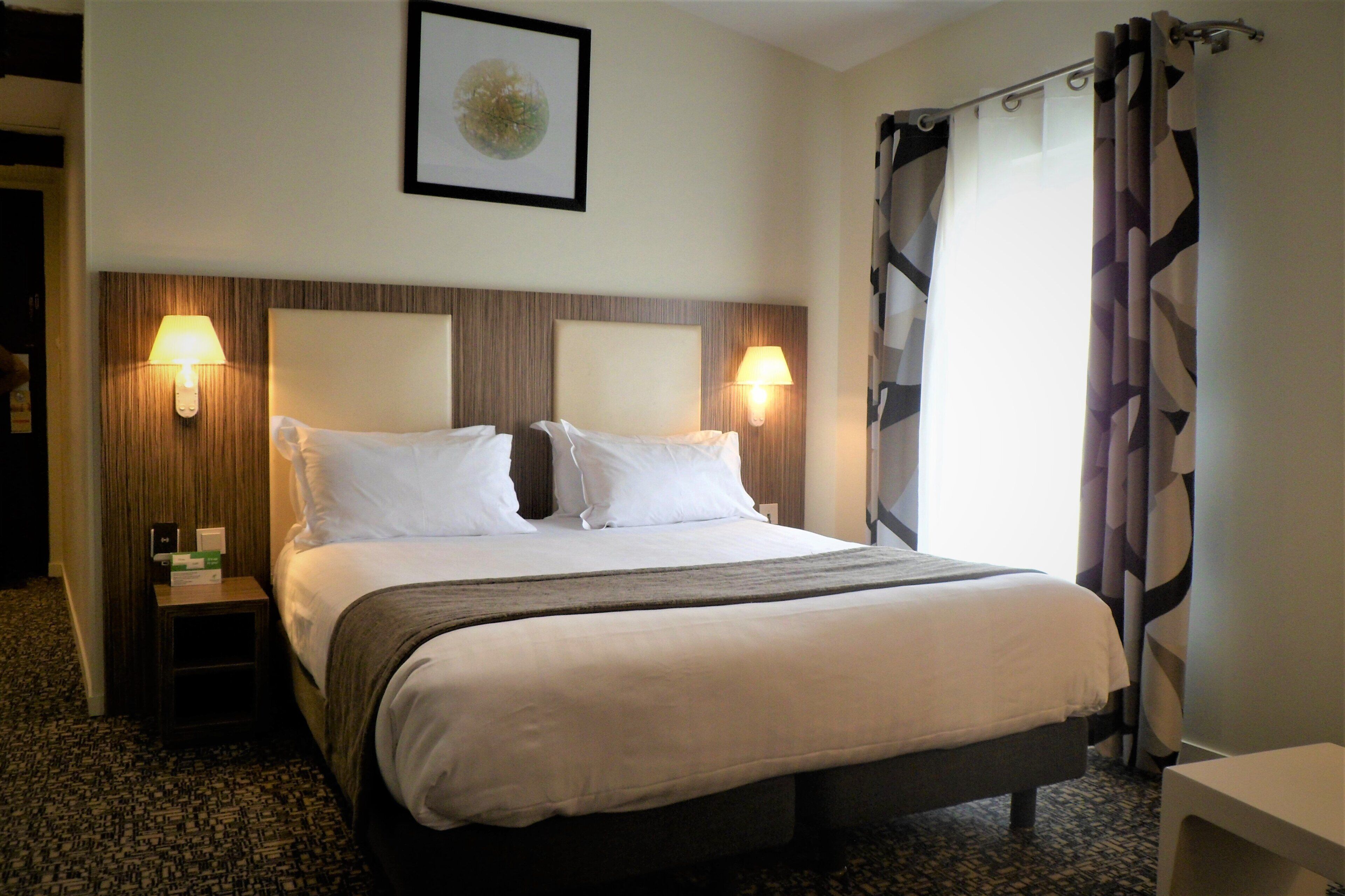 A hotel room with a large bed, two bedside lamps, a framed picture on the wall, and a window with curtains.