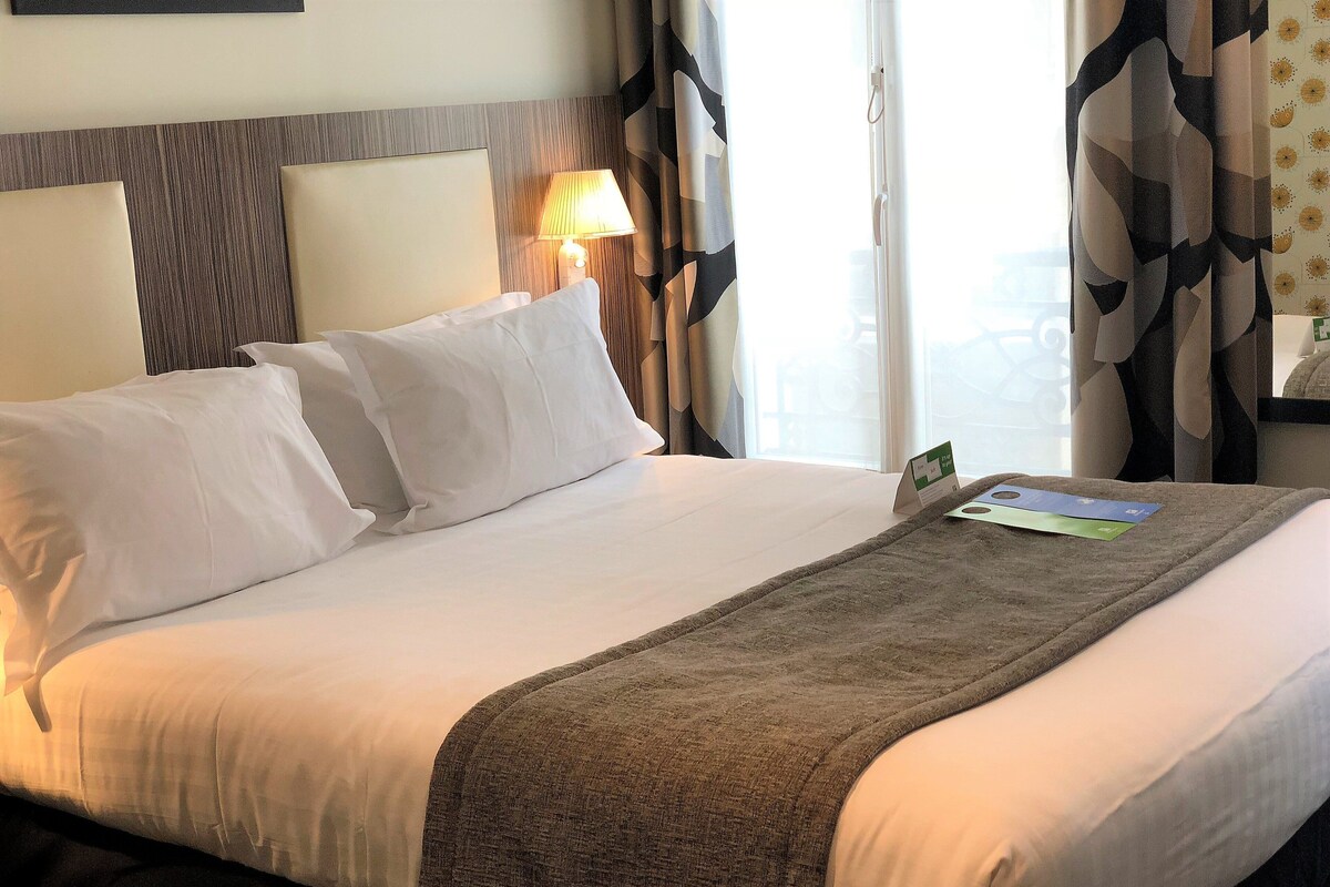Quarto de hotel com uma cama grande, roupa de cama branca, manta marrom, mesa de cabeceira com abajur e janela com cortinas.